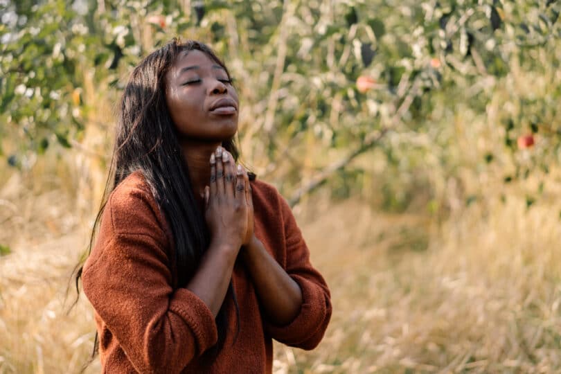 Mulher orando com os olhos fechados e as mãos juntas, ao ar livre.
