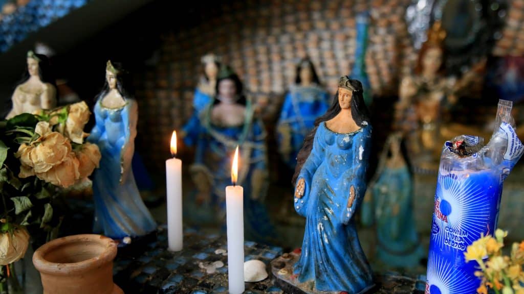 Estátuas dispersas da deusa Iemanjá. Algumas velas acesas e, à direita, uma vela azul.