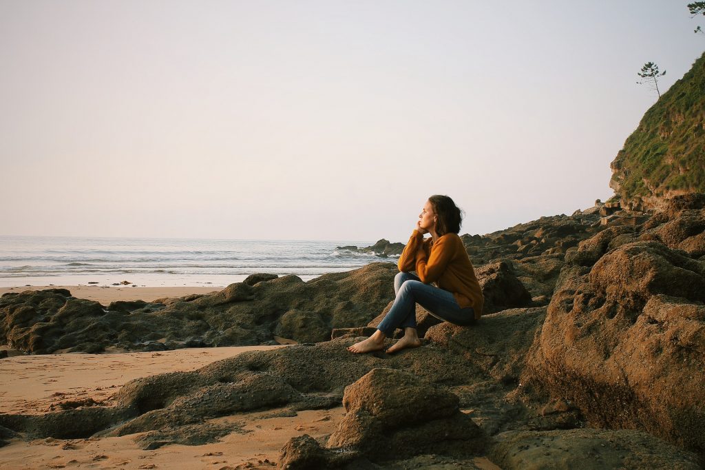 Mulher branca sentada em pedras numa praia.