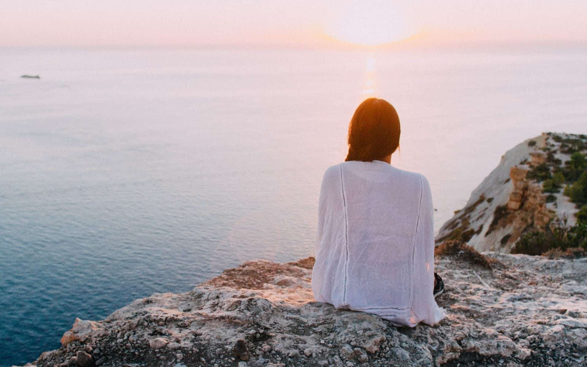 Uma mulher contemplando, de uma montanha, um horizonte com sol e um grande mar.