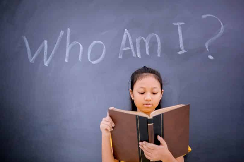 Uma garota lendo um livro. No fundo, situado numa lousa, os dizeres "Who am I?"