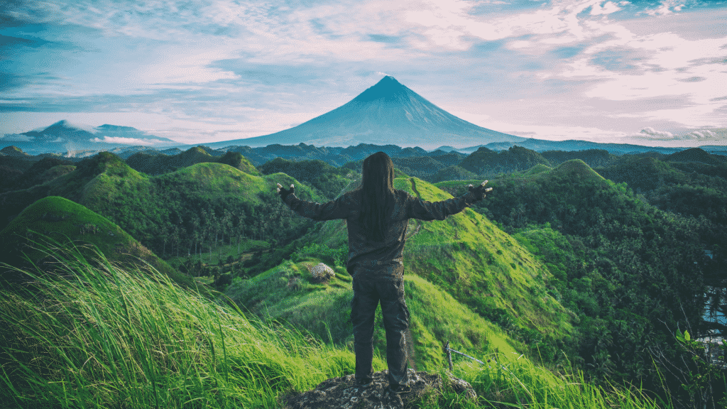 Uma mulher de braços abertos observando vários cumes verdes e uma montanha, ao fundo.
