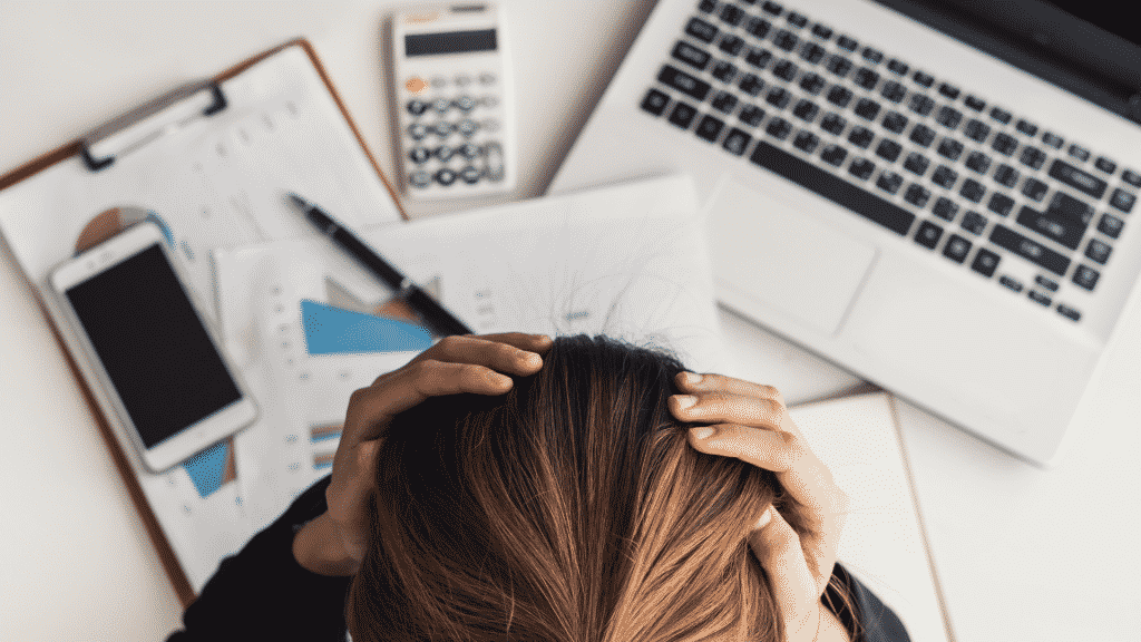 Mulher com as duas mãos na cabeça em frente ao seu celular, notebook, calculadora e alguns papéis