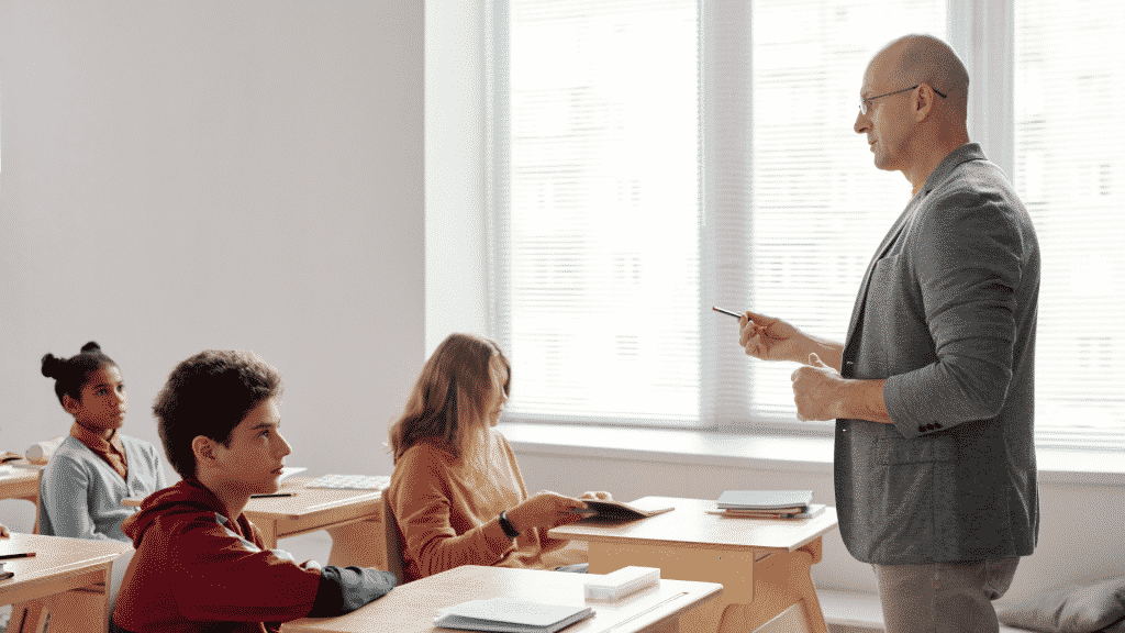 Professor em frente da sala, com alunos prestando atenção a sua fala