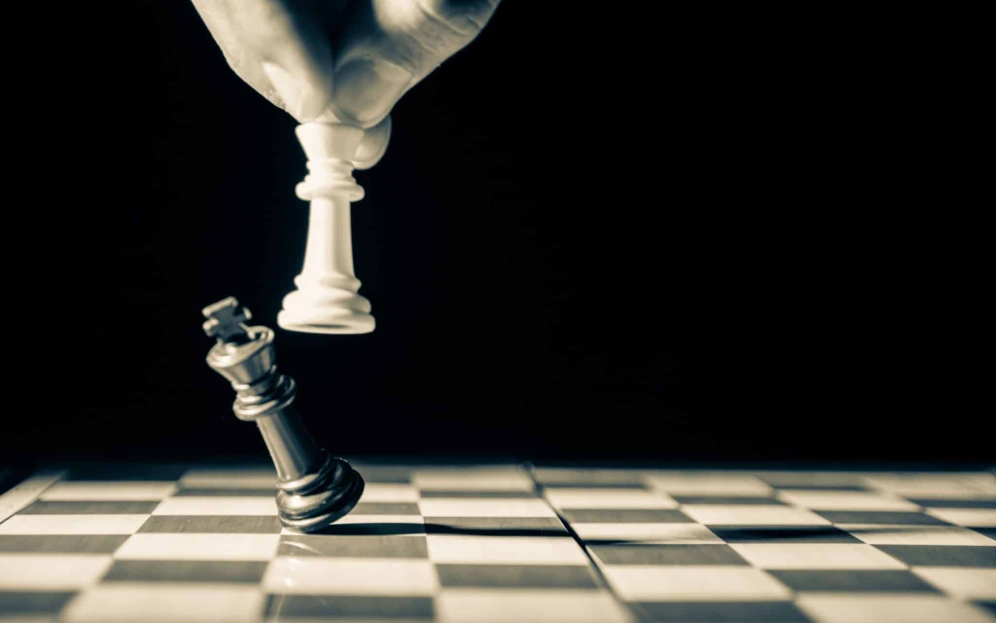 Duas peças de xadrez. Um homem manipula uma - branca - que derruba uma preta.
