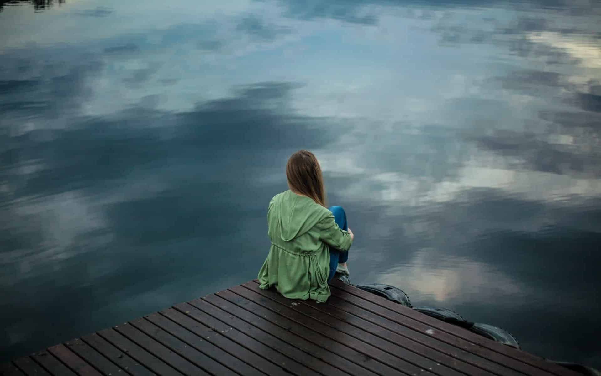 Uma mulher de casaco verde sentando próxima à borda de um rio.