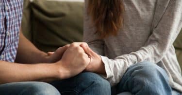 Duas pessoas segurando as mãos. Elas estão sentadas num sofá.