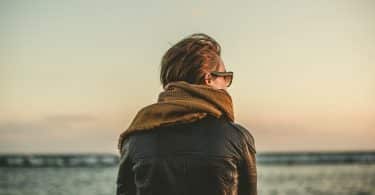 Homem branco de costas usando roupas de frio na praia.