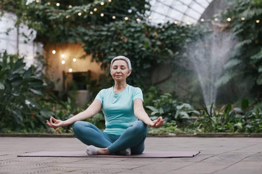 Mulher branca sentada numa área externa meditando.