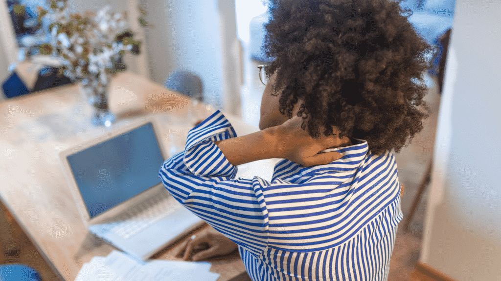 Mulher sentada na frente do notebook com a mão no pescoço, em sinal de dor