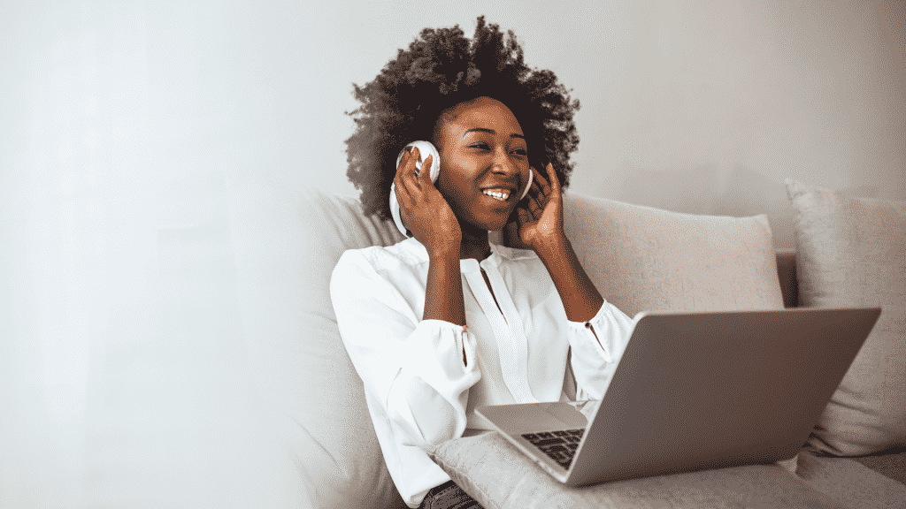 Mulher sentada em um sofá ouvindo música, sorrindo e com um notebook no colo