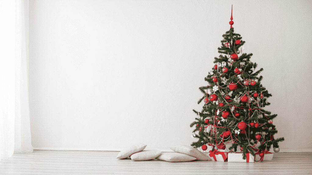 Uma árvore-de-natal. Embaixo dela, caixas de presentes natalinos. À esquerda, almofadas brancas.