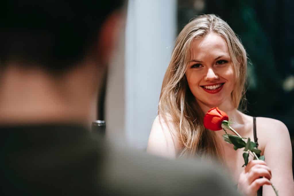 Uma mulher loira sorrindo, em segundo plano. Ela segura uma rosa. Em primeiro plano, desfocado, um homem.