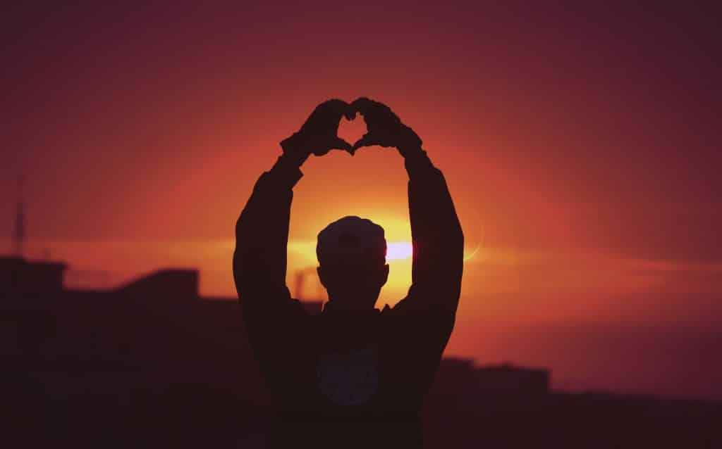 Silhueta de homem com as mãos formando coração no pôr do sol.