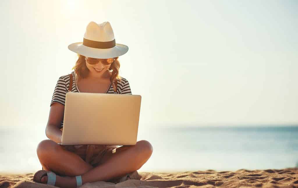 Uma mulher usando um notebook. Ela senta na areia e está sorrindo.