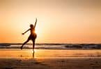 Uma mulher dançando numa praia que tem um pôr do sol.