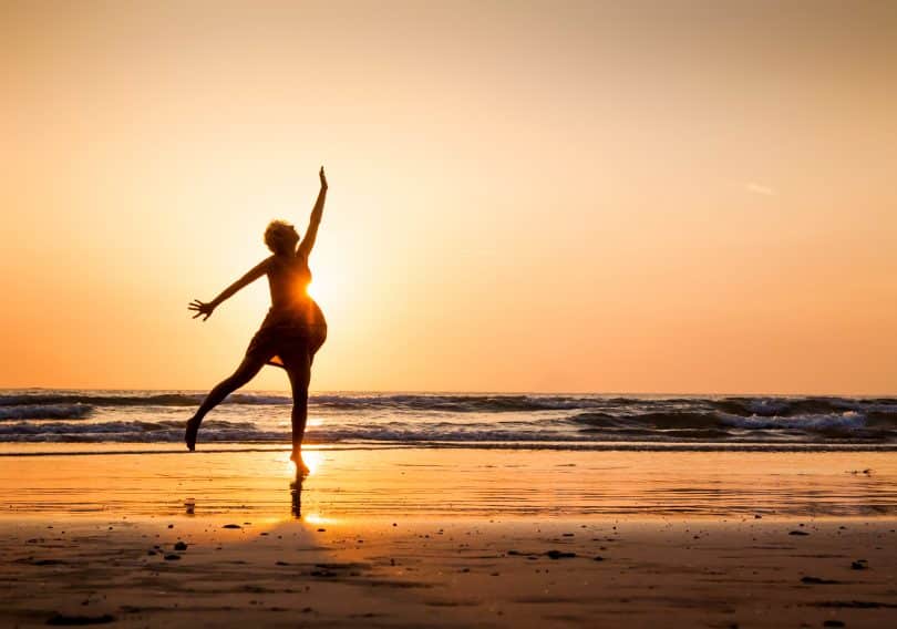 Uma mulher dançando numa praia que tem um pôr do sol.