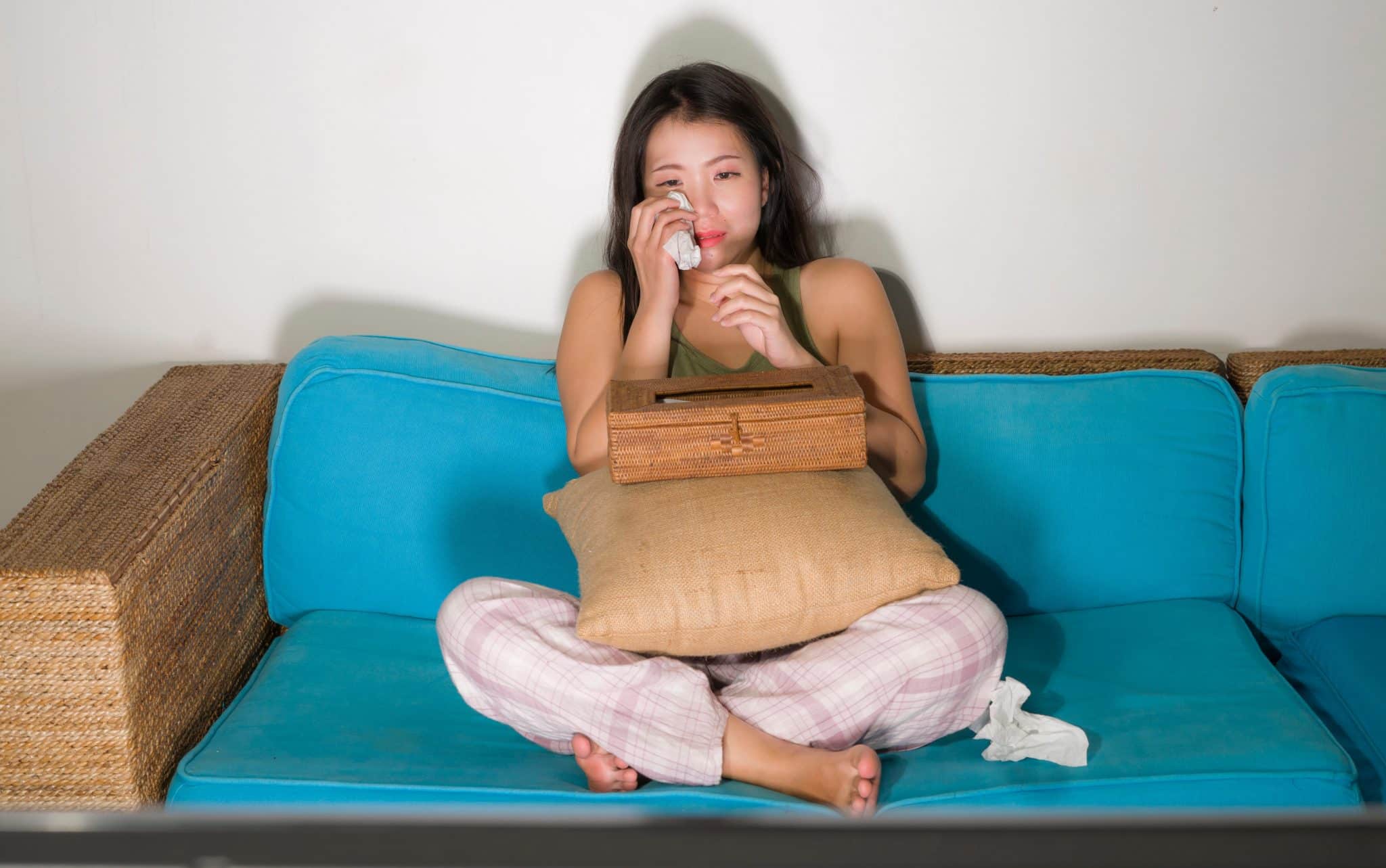 Uma mulher asiática chorando. Ela está sentada num sofá e tem consigo uma caixa de lenços de papel.