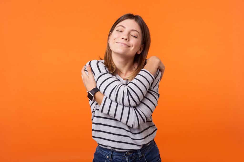 Mulher com cabelo castanho em camisa de manga comprida em pé com os olhos fechados se abraçando,