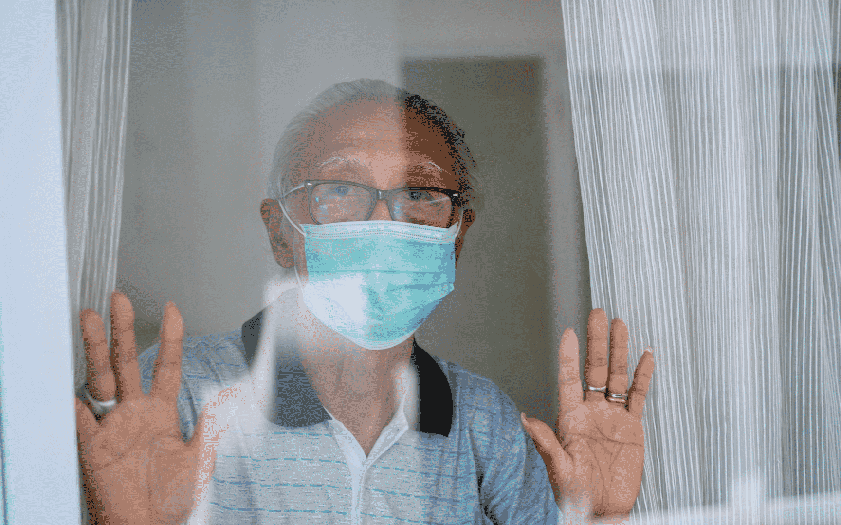 Um idoso trajando uma máscara no seu rosto e olhando através de uma janela.