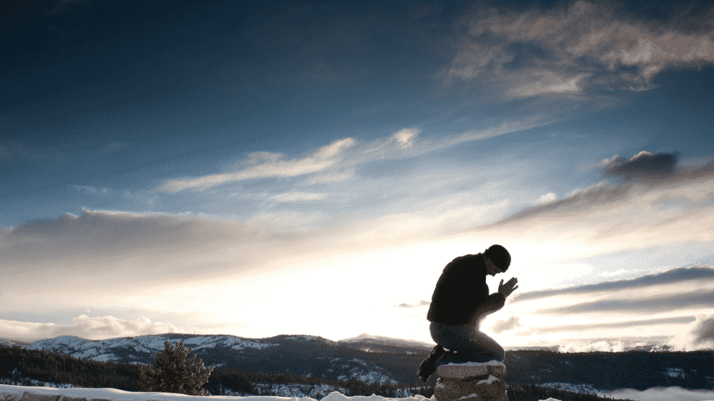 Homem de joelhos em cima de uma pedra, em sinal de oração. Ao fundo, uma paisagem com nuvens e montanhas