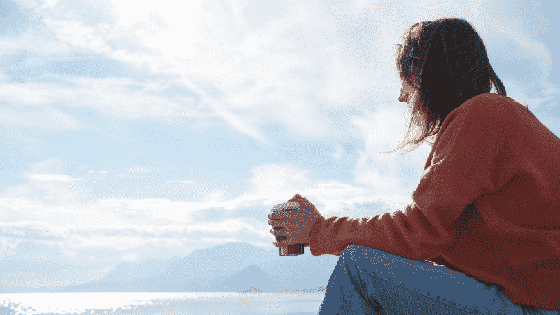 Menina sentada com um copo na mão, observando o mar