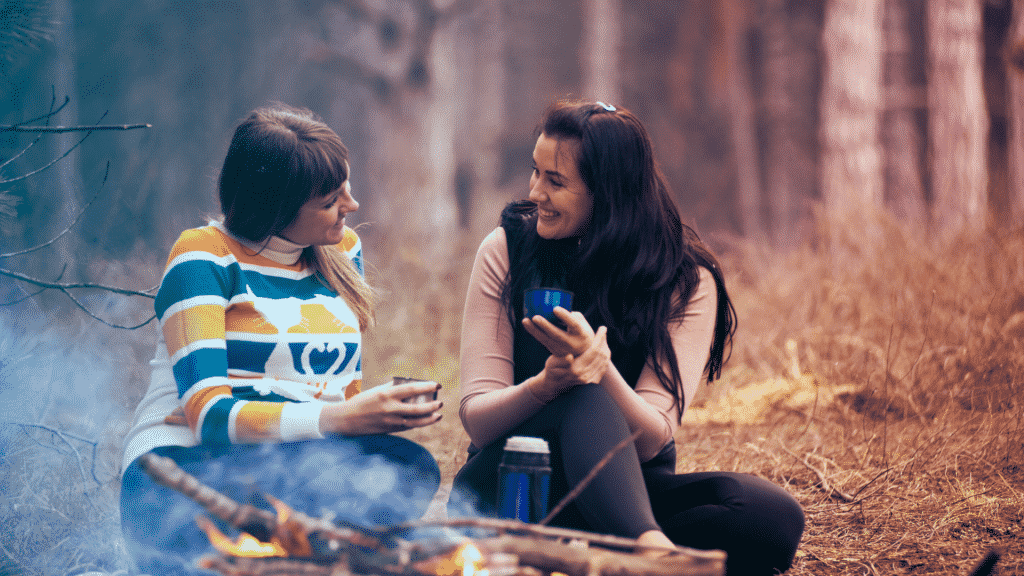 Duas amigas sentadas na frente de uma fogueira, sorrindo e conversando