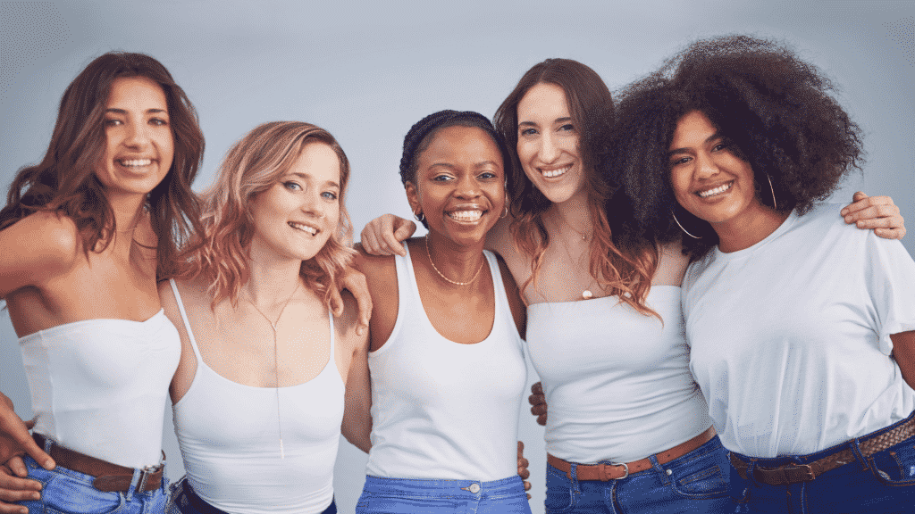 Mulheres de várias etnias se abraçando e sorrindo. Todas estão de blusa branca e calça jeans