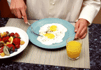 Pessoa se alimentando de ovos fritos. Na mesa há também um copo de suco de laranja e um prato com frutas diversas