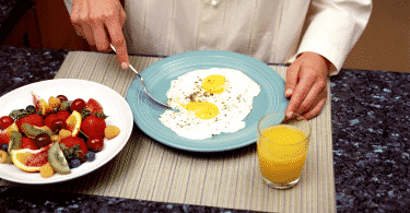 Pessoa se alimentando de ovos fritos. Na mesa há também um copo de suco de laranja e um prato com frutas diversas
