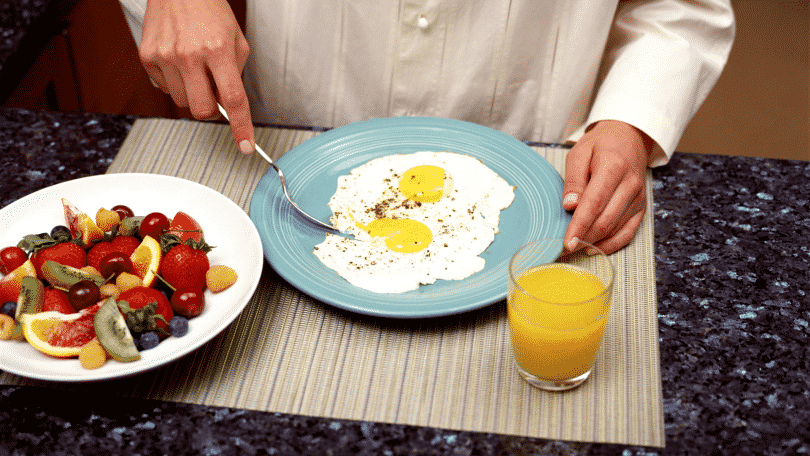 Pessoa se alimentando de ovos fritos. Na mesa há também um copo de suco de laranja e um prato com frutas diversas