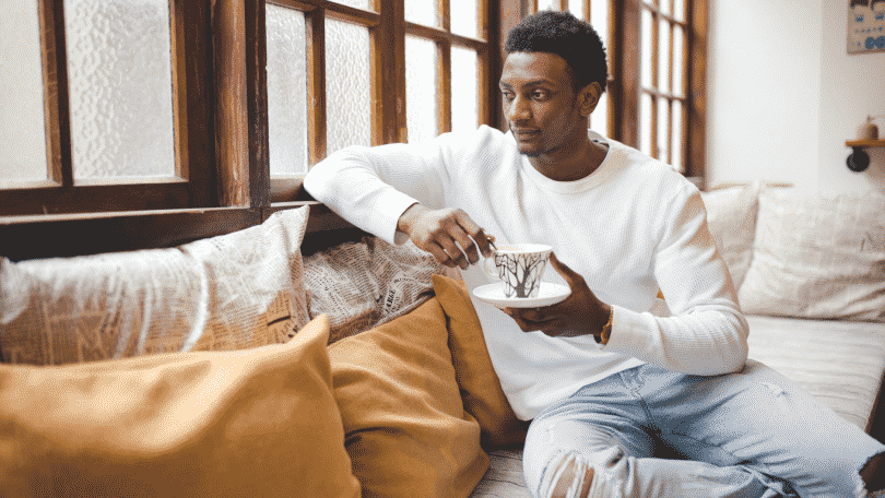 Homem sentado em um sofá, tomando chá e olhando para a janela