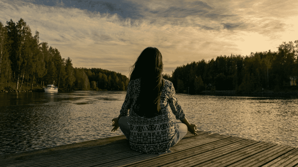 Uma mulher meditando em frente a um lago.