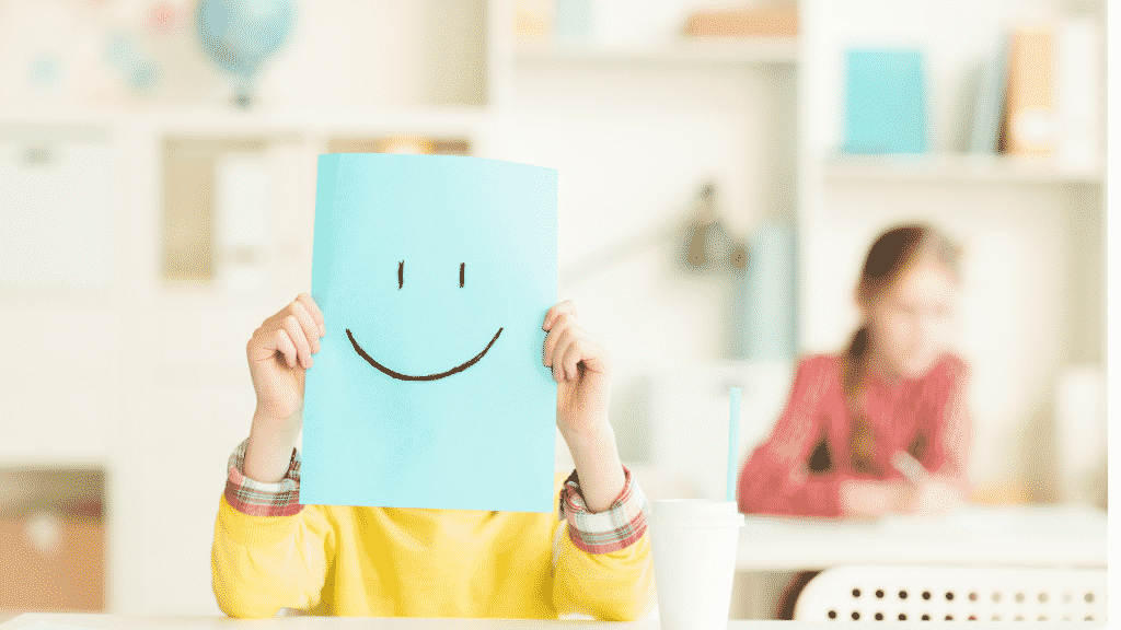 Uma pessoa segurando uma folha de papel sulfite azul em frente do seu rosto. Neste papel, um desenho que representa uma feição/rosto feliz.