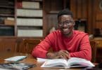 Homem negro sentado em uma biblioteca. Ele sorri. Há um livro a sua frente.