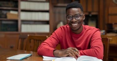 Homem negro sentado em uma biblioteca. Ele sorri. Há um livro a sua frente.