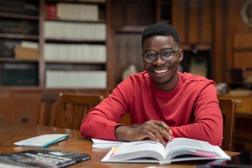 Homem negro sentado em uma biblioteca. Ele sorri. Há um livro a sua frente.