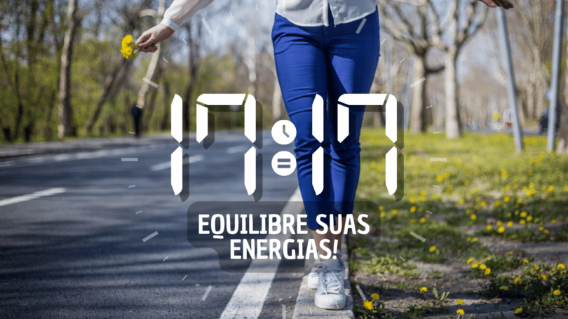 Hora igual 17:17 e texto "Equilibre suas energias!" aplicado por cima de foto das pernas de uma mulher que veste uma calça azul. Ela se equilibra na guia de uma calçada que está coberta de gramas e de flores amarelas. Ela segura pequenas flores amarelas em uma mão. Ao fundo, há uma rua e árvores.