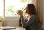Mulher tomando café e olhando por uma janela.