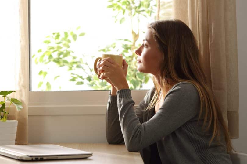 Mulher tomando café e olhando por uma janela.