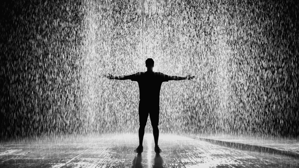 Um homem, de braços abertos, tomando banho de chuva.