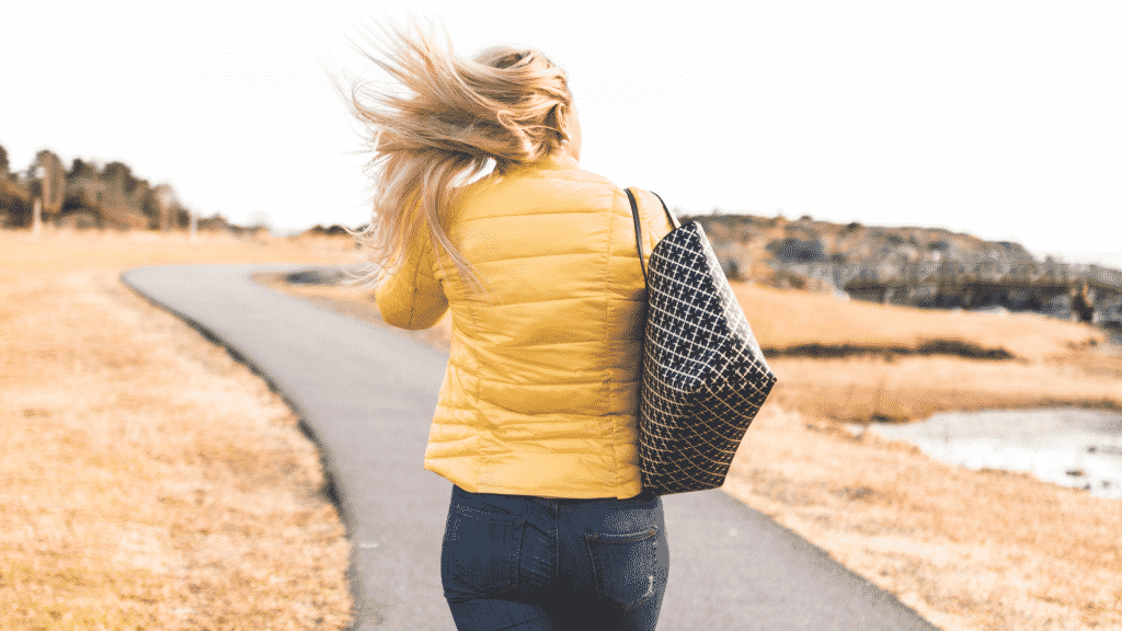 Mulher andado por um caminho carregando uma bolsa e com os cabelos ao vento