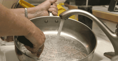 Pessoa lavando uma panela na pia. A torneira está ligada