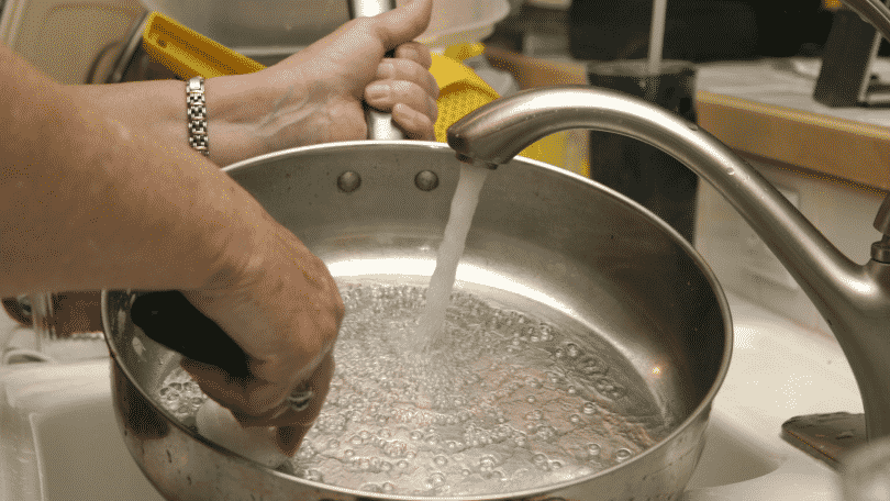 Pessoa lavando uma panela na pia. A torneira está ligada
