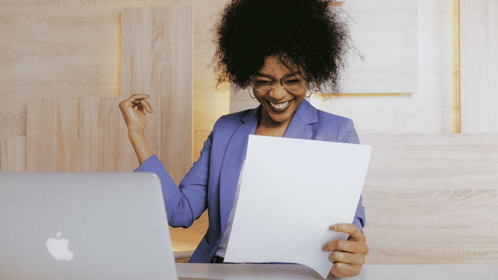 Mulher sorrindo com um papel na mão e um notebook na sua frente