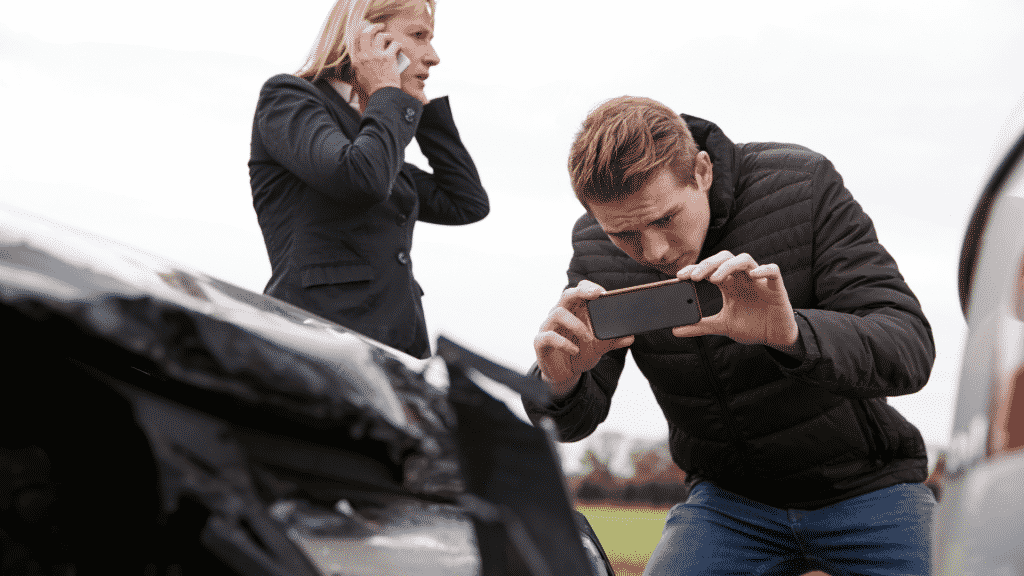 Um homem realizando capturas fotográficas de um carro batido. Ao seu lado esquerdo, uma mulher realizando uma ligação via telefone celular.