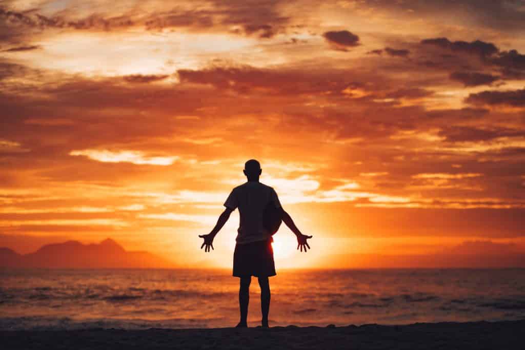 Homem de braços abertos em frente ao mar durante um por do sol.