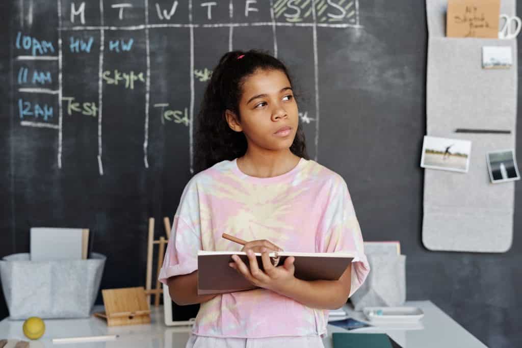 Menina com um caderno na mão. Ela está olhando para o lado com um semblante pensativo.