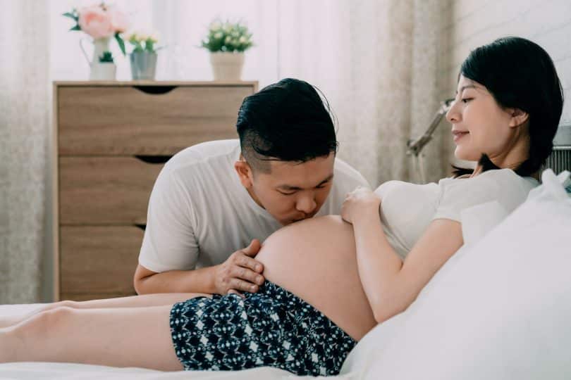 Uma mulher grávida e, ao seu lado, um homem beijando sua barriga.