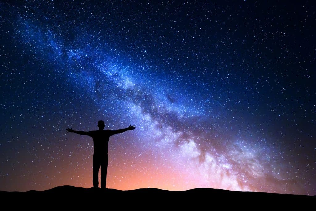 Silhueta de homem de braços abertos frente a um céu colorido e estrelado à noite